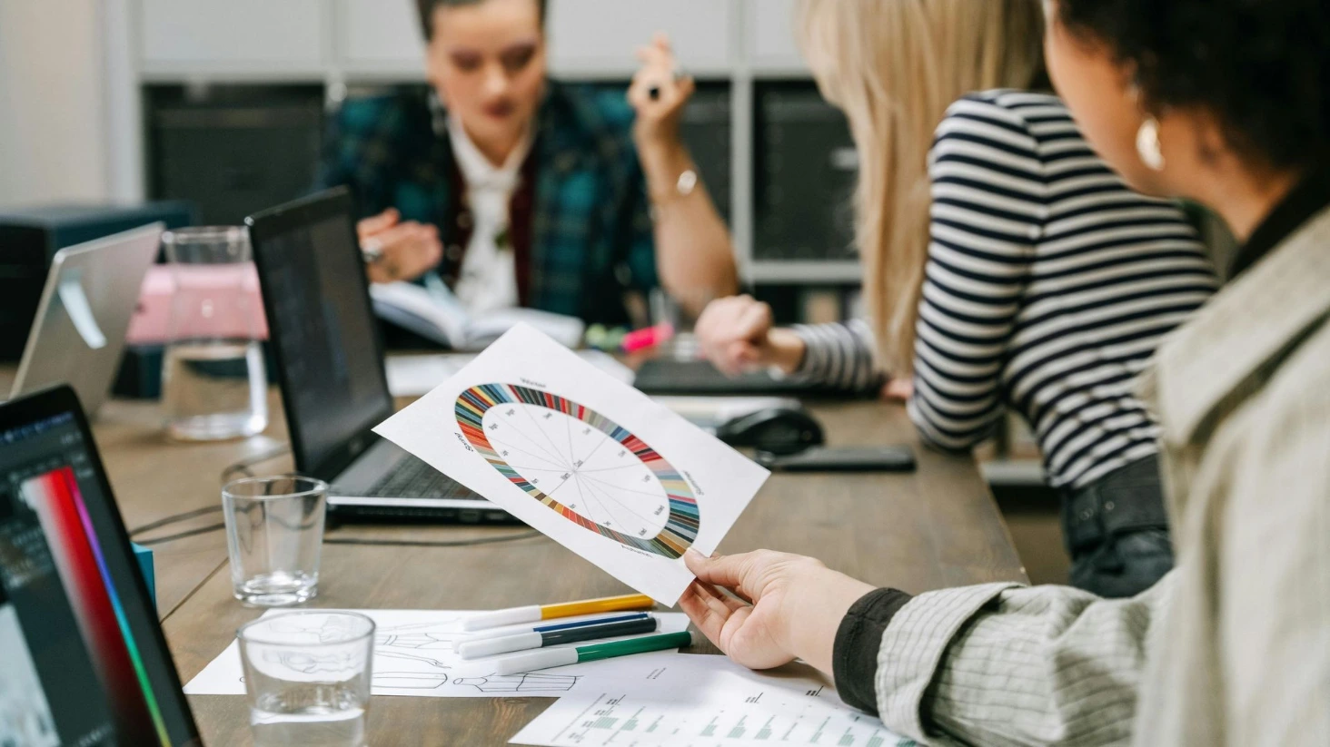 Réunion créative : main présentant une roue chromatique sur papier. Équipe collaborant sur ordinateurs, illustrant la phase de conception stratégique.