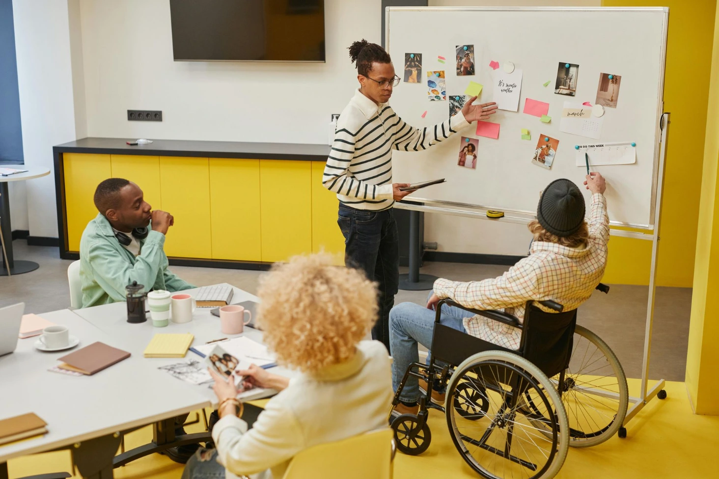 Séance de brainstorming inclusive : un collaborateur explique des notes au tableau devant une équipe diversifiée, dont un employé en fauteuil roulant.