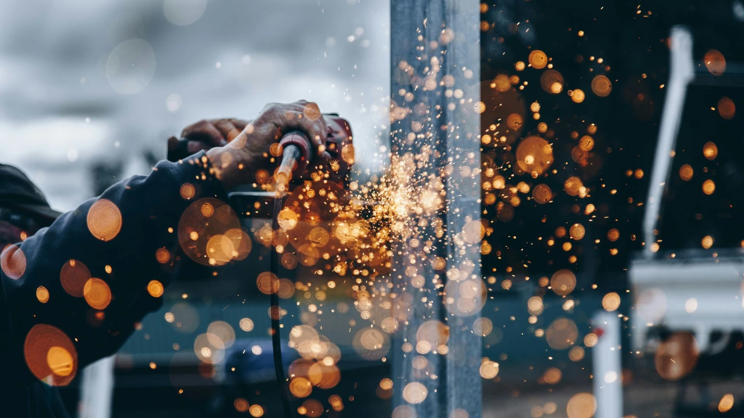 Gros plan sur les mains d'un ouvrier du bâtiment utilisant une meuleuse d'angle sur une poutre en métal, générant une importante gerbe d'étincelles lumineuses et dorées sur un chantier